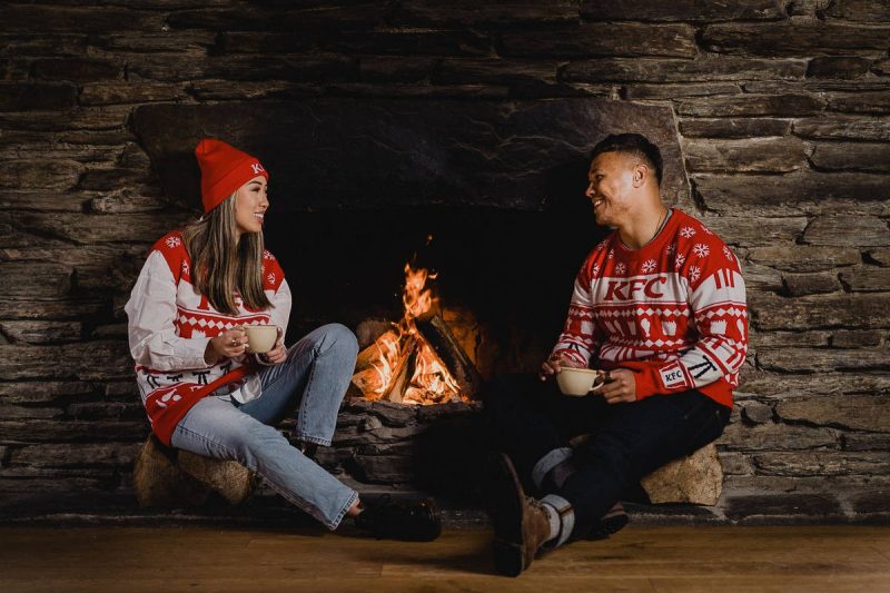 Two people sitting in a KFC vest and sweater in front of a fireplace